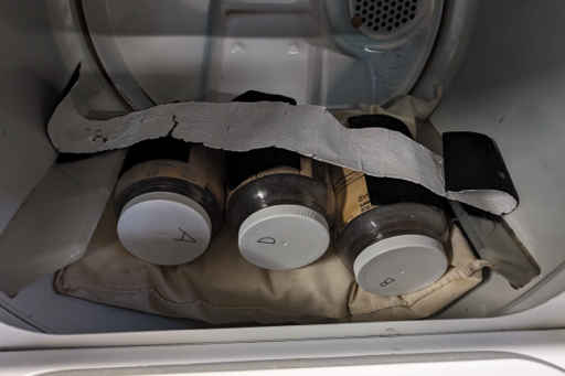 Three jugs placed inside a dryer. They are supported by a pillow, and held in place by a long wide strip of velcro. The velcro is attached to the dryer fins and the jars. The jar lids are labeled A, B and D.