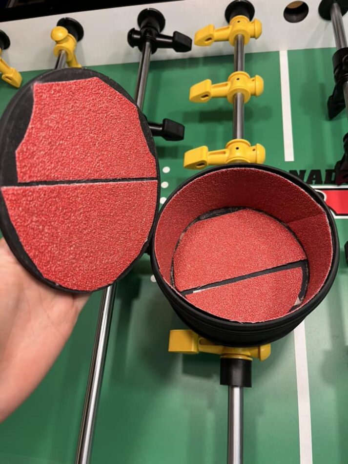 An open rock tumbler canister, several inches wide and tall, lined with bright red sandpaper. The tumbler is positioned over a green tornado foosball playing field.